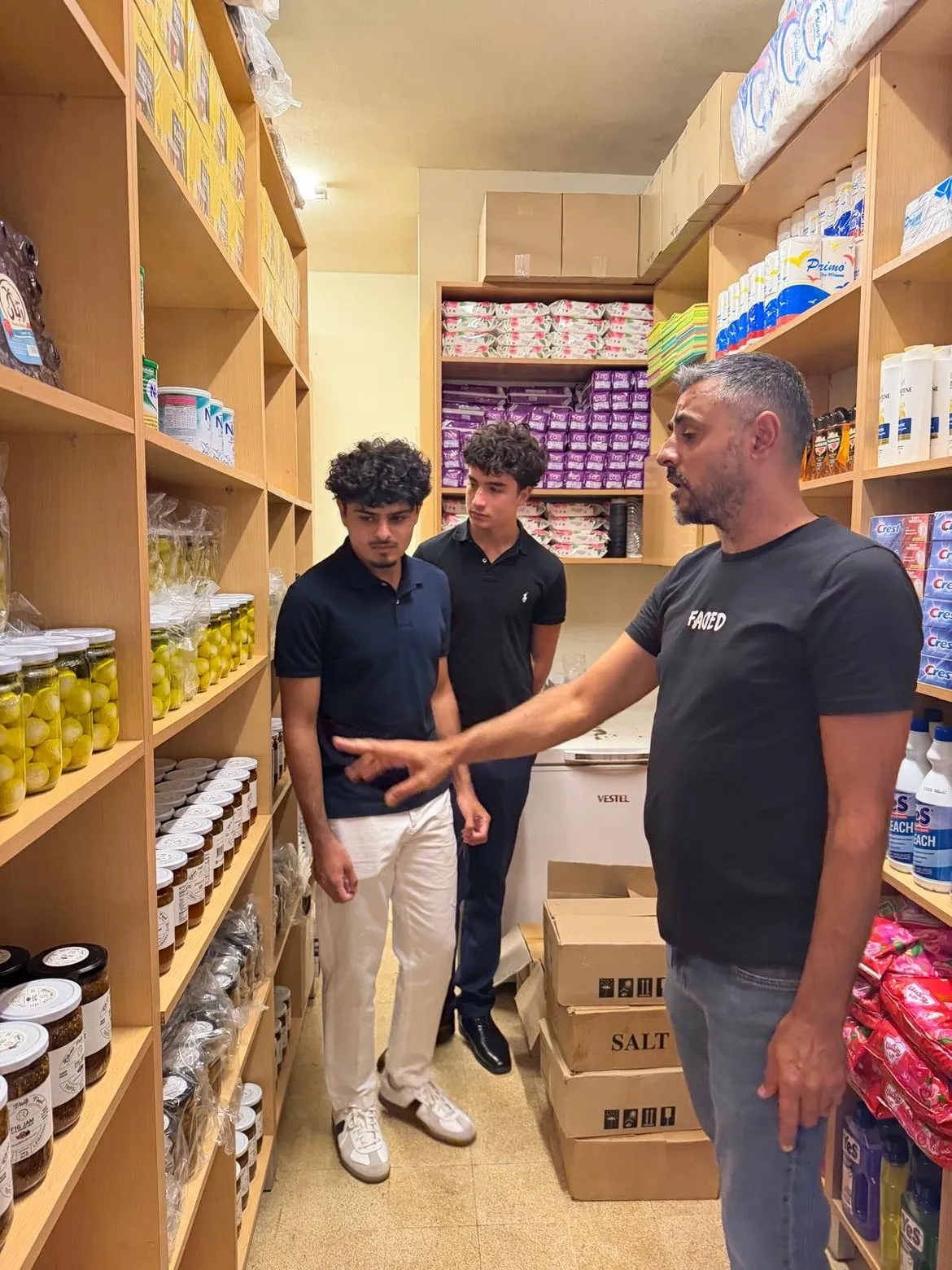 A Beit el Baraka team member shows two students shelves of jars and cleaning supplies.