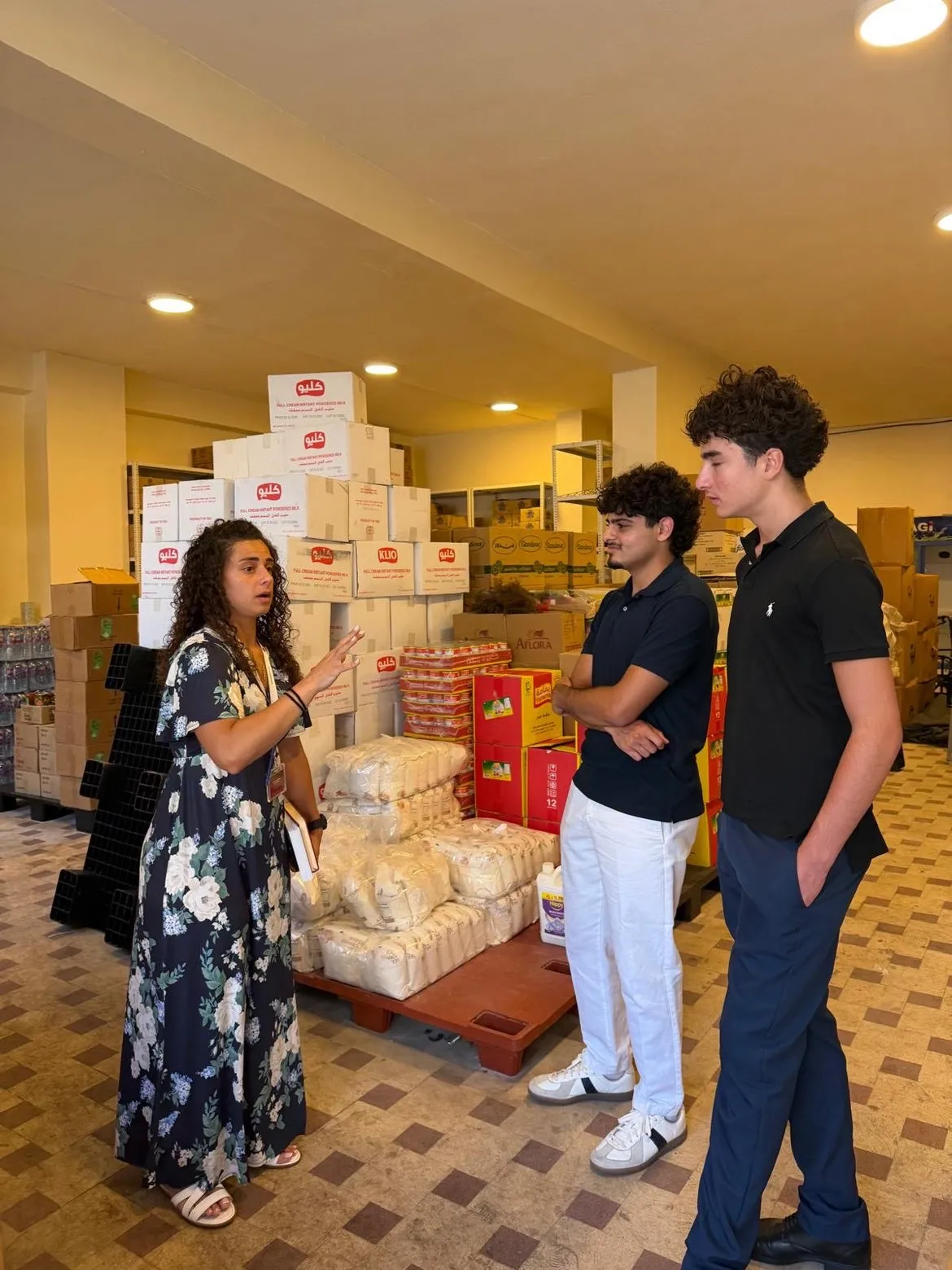 A Beit el Baraka team member talks with two students in a hall of stacked boxes of food.
