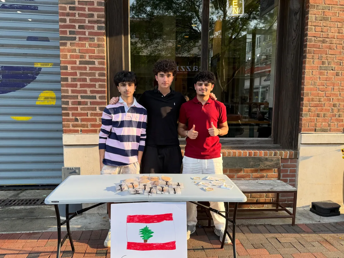 Three students at the Bethesda bake sale with cookies and bracelets on the table and a hand-drawn Lebanese flag.
